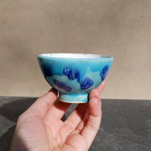 May include: A small, handmade ceramic bowl with a turquoise glaze. The bowl features dark blue and purple abstract designs. The interior is white. The bowl is held in a hand, showcasing its size and shape. The bowl is a decorative item.