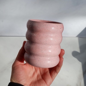 Może przedstawiać: Różowy ceramiczny kubek o unikalnym, teksturowanym wzorze. Kubek ma szereg zaokrąglonych, ułożonych w stos żeber i jest nakrapiany ciemnymi plamkami. Kubek jest trzymany w dłoni, prezentując jego kształt i rozmiar.