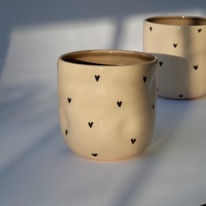 Puede incluir: Dos tazas de cerámica de color crema con patrones de corazones negros. Las tazas son de formas y tamaños ligeramente diferentes.