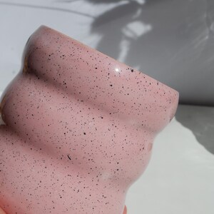 May include: A light pink ceramic mug with a speckled finish featuring small black dots throughout the surface. The mug has a distinct wavy, rippled texture along its exterior body, creating a series of horizontal ridges from the rim to the base.
