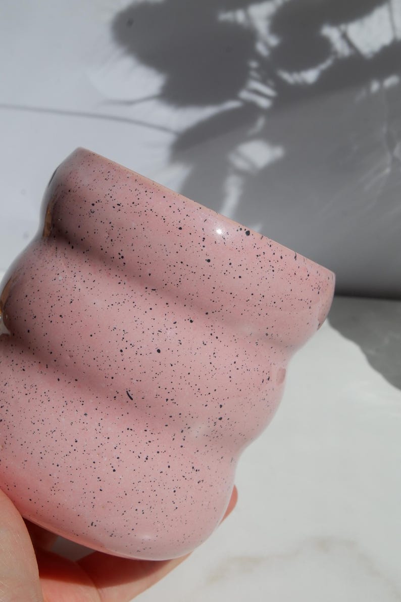 May include: A light pink ceramic mug with a speckled finish featuring small black dots throughout the surface. The mug has a distinct wavy, rippled texture along its exterior body, creating a series of horizontal ridges from the rim to the base.