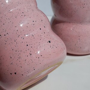 May include: Two pink ceramic mugs with a speckled black pattern. Each mug features a rounded, bulbous shape with a smooth, glossy finish and a visible unglazed beige base rim. The mugs are positioned close together, showing their curved design.