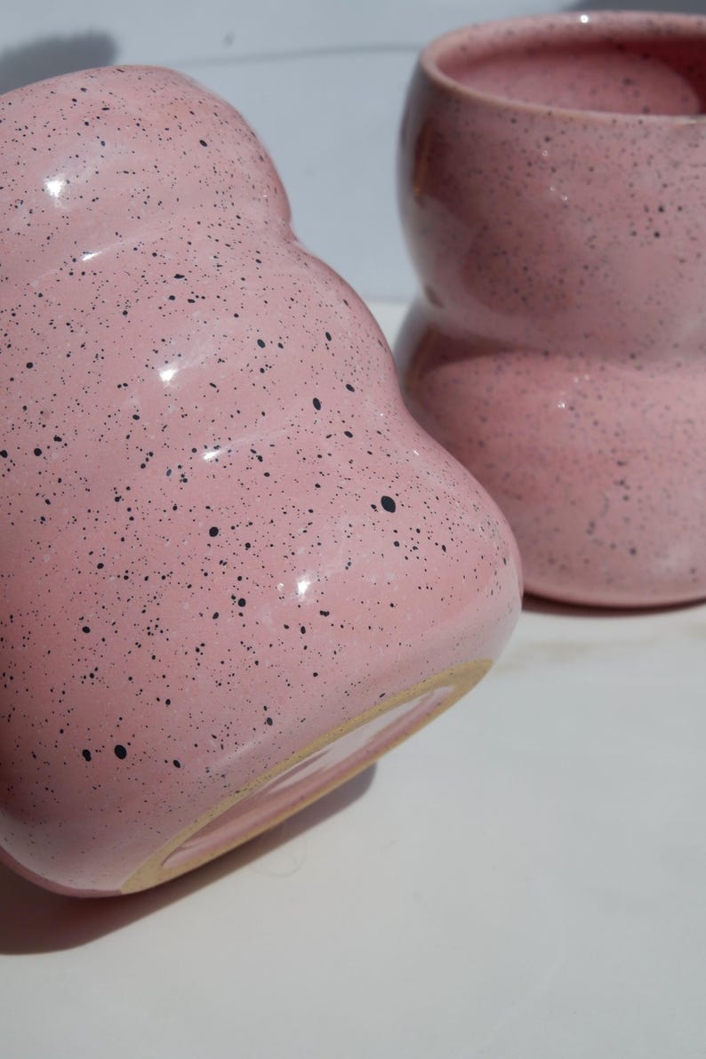 May include: Two pink ceramic mugs with a speckled black pattern. Each mug features a rounded, bulbous shape with a smooth, glossy finish and a visible unglazed beige base rim. The mugs are positioned close together, showing their curved design.