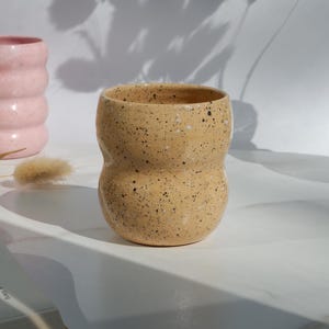 May include: A speckled, beige ceramic tumbler with a unique, curved shape. The handmade cup has a wide opening and a rounded base. A pink, ribbed ceramic cup is visible in the background. The tumbler is likely used for drinking beverages.