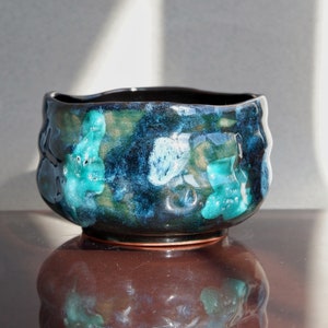 May include: A dark blue ceramic bowl with a reactive glaze featuring green and blue abstract patterns. The bowl has a slightly uneven rim and a rounded base.