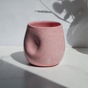 May include: A rounded ceramic cup in a soft pink hue with a speckled finish. The cup features a distinct, concave thumb indentation on its side, creating a unique, ergonomic shape. The surface is smooth, and the rim is circular and open.