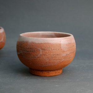 May include: A small, round, terracotta-colored ceramic bowl with a white glaze on the rim. The bowl has a slightly textured surface and a wide, rounded base.