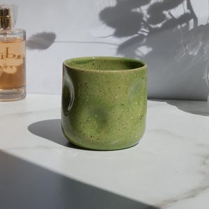 Puede incluir: Una pequeña taza de cerámica cilíndrica con un esmalte verde claro moteado. La superficie exterior presenta varias hendiduras sutiles y redondeadas presionadas en la arcilla, creando una textura táctil y orgánica alrededor del cuerpo.