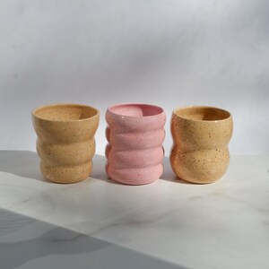 May include: Three ceramic cups with a wavy, ribbed silhouette and a speckled finish. Two cups are a light tan color, while the center cup is a soft pastel pink. Each cup features a rounded, textured body with a smooth rim and a wide opening.