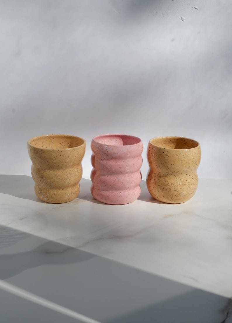 May include: Three ceramic cups with a wavy, ribbed silhouette and a speckled finish. Two cups are a light tan color, while the center cup is a soft pastel pink. Each cup features a rounded, textured body with a smooth rim and a wide opening.
