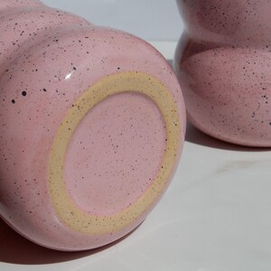 May include: Two pink ceramic mugs with a speckled finish and a rounded, wavy silhouette. One mug is positioned horizontally to show its circular base with a light tan ring, while the other stands upright, displaying its curved, bulbous shape.