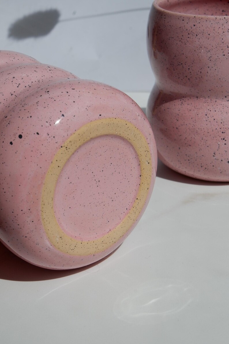 May include: Two pink ceramic mugs with a speckled finish and a rounded, wavy silhouette. One mug is positioned horizontally to show its circular base with a light tan ring, while the other stands upright, displaying its curved, bulbous shape.
