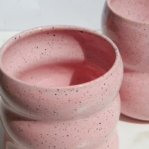 May include: Two pink ceramic cups with a speckled black pattern. Each cup features a rounded, ribbed, or tiered cylindrical shape with a wide opening at the top. The surface has a smooth finish with visible dark spots distributed across the pink glaze.