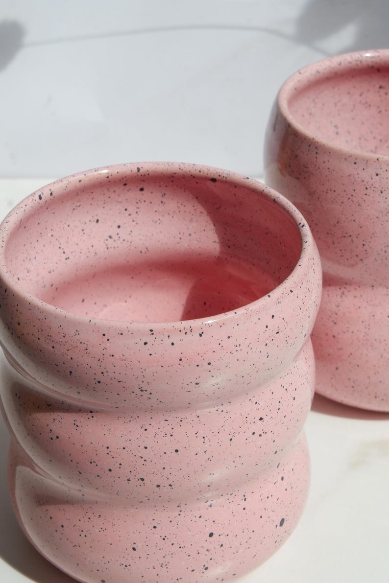 May include: Two pink ceramic cups with a speckled black pattern. Each cup features a rounded, ribbed, or tiered cylindrical shape with a wide opening at the top. The surface has a smooth finish with visible dark spots distributed across the pink glaze.