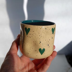 May include: A handmade ceramic mug with a beige speckled exterior and a teal interior. The mug features three green heart designs. The mug is held in a hand.