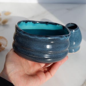 May include: A handmade ceramic matcha bowl and a small matching vessel. The bowl is a deep blue with a turquoise interior and a textured, ridged exterior. The small vessel is also blue. The items are designed for preparing and serving matcha tea.