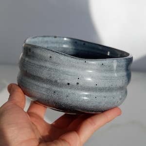 May include: A handmade ceramic bowl with a textured, ridged exterior. The bowl is a gradient of gray and blue hues, speckled with dark spots. The bowl is held in a hand, showcasing its unique shape and design.