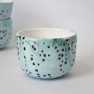 May include: A light blue ceramic bowl with a speckled pattern of black and gold dots. The bowl is handmade and has a unique, organic shape.