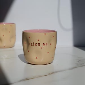以下が含まれることがあります： 赤いハートのアクセントと「I LIKE ME」の文字が入ったベージュ色のセラミックマグ。マグの内側はピンク色です。マグは少し不規則な形をしており、白い表面に置かれています。