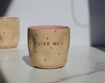 May include: Beige ceramic mug with red heart accents and the text "I LIKE ME". The interior of the mug is pink. The mug has a slightly irregular shape and is sitting on a white surface.
