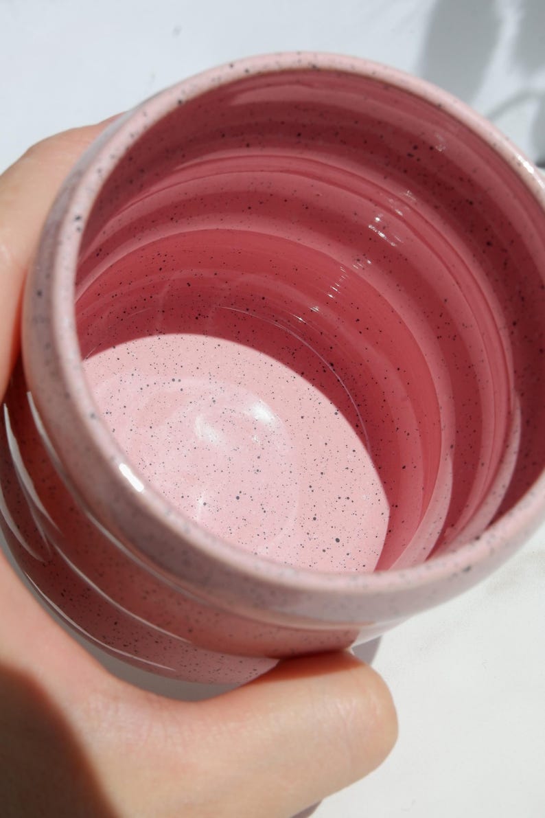 May include: A cylindrical ceramic mug with a glossy light pink glaze and dark speckles throughout the interior and exterior. The mug features horizontal ridges along the outer wall and a smooth, rounded rim, showcasing a speckled finish.