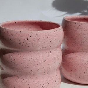 May include: Two ceramic mugs with a light pink glaze and dark speckles. Each mug features a distinct wavy, rippled silhouette with three horizontal rounded ridges along the body, creating a textured appearance with a smooth, glossy finish.