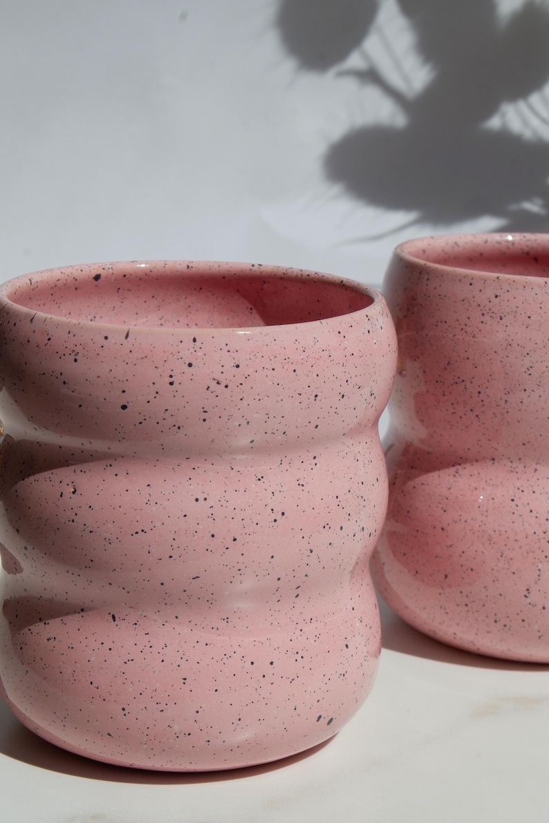 May include: Two ceramic mugs with a light pink glaze and dark speckles. Each mug features a distinct wavy, rippled silhouette with three horizontal rounded ridges along the body, creating a textured appearance with a smooth, glossy finish.