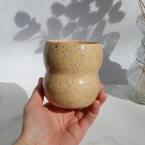 May include: A speckled, beige ceramic cup with a unique, curved shape. The cup has a wide opening and a rounded base. The surface is covered in small, dark brown speckles. The cup is held in a hand, with a white background and shadows.