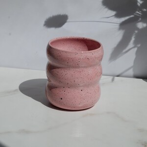 May include: A cylindrical ceramic cup with a light pink glaze and dark speckles throughout. The vessel features three distinct horizontal ridges or indentations along its body, creating a wavy, rounded profile with an open top and a flat base.
