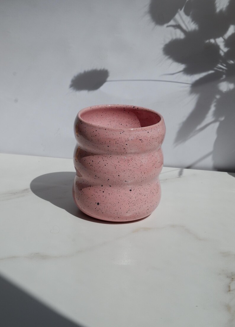 May include: A cylindrical ceramic cup with a light pink glaze and dark speckles throughout. The vessel features three distinct horizontal ridges or indentations along its body, creating a wavy, rounded profile with an open top and a flat base.