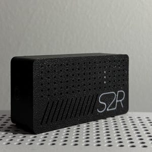 Puede incluir: Un dispositivo rectangular negro con una superficie texturizada y el texto blanco "S2R". El dispositivo tiene una serie de pequeños agujeros y líneas anguladas en la parte delantera. El objeto está sobre una superficie perforada.