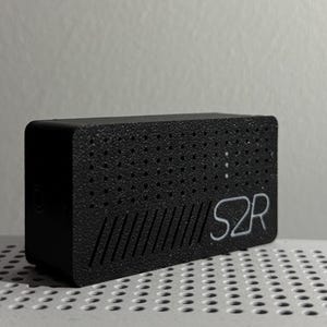 Puede incluir: Un dispositivo rectangular negro con una superficie texturizada. El dispositivo tiene pequeños agujeros y líneas diagonales en la parte delantera. Las letras "S2R" están impresas en blanco en el lateral.