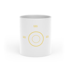 Könnte beinhalten: Ein weißer Keramikbecher mit einem gelben Grafikdesign auf der Vorderseite. Das Design besteht aus zwei konzentrischen Kreisen in der Mitte des Bechers, umgeben von der Zahl 111, die in Gelb oben, links und rechts neben den Kreisen aufgedruckt ist.