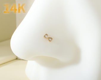 May include: A gold nose ring with a small bow design and two clear stones. The ring is labeled "14K".