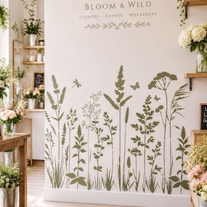 May include: A wall decal featuring olive green botanical silhouettes of wildflowers, grasses, and insects on a white wall. The text reads BLOOM & WILD FLOWERS EVENTS WORKSHOPS with a decorative floral flourish centered above the plant designs.