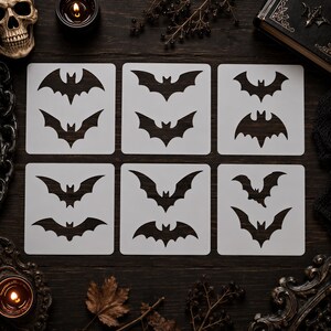 May include: A set of six square white stencils, each featuring two distinct black bat silhouettes. The stencils are arranged in a grid pattern, displaying various wing shapes and flight positions for the bats, providing a clear template for decorative applications.