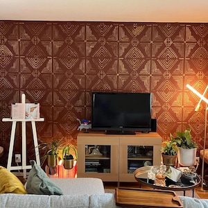 May include: A wall covered in deep red wallpaper featuring a repeating gold geometric diamond pattern. The wall serves as a backdrop for a black television set on a light wood media console, a white wooden easel, and various indoor potted plants.