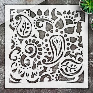May include: A square white stencil featuring a detailed cutout pattern of paisley shapes, swirling floral motifs, and small circular accents. The design is symmetrical and intricate, with negative space creating the complex decorative pattern.