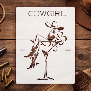 May include: A rectangular white sign featuring a dark brown graphic of a person in a cowboy hat and boots sitting inside a large martini glass. The text COWGIRL is printed at the top, with EST 1889 on either side of the central graphic illustration.
