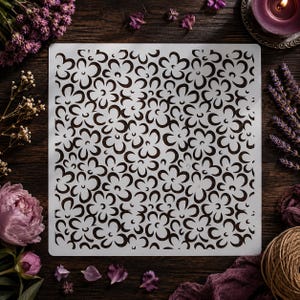 May include: A white floral stencil with a repeating pattern of stylized flowers. The stencil is square and sits on a dark wooden surface, surrounded by purple flowers and a candle.