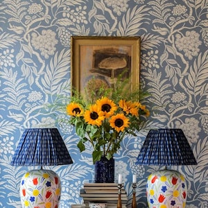 a table with two blue lamps, a vase of yellow sunflowers, and a framed picture on the wall. The table also has a stack of books and some decorative items.