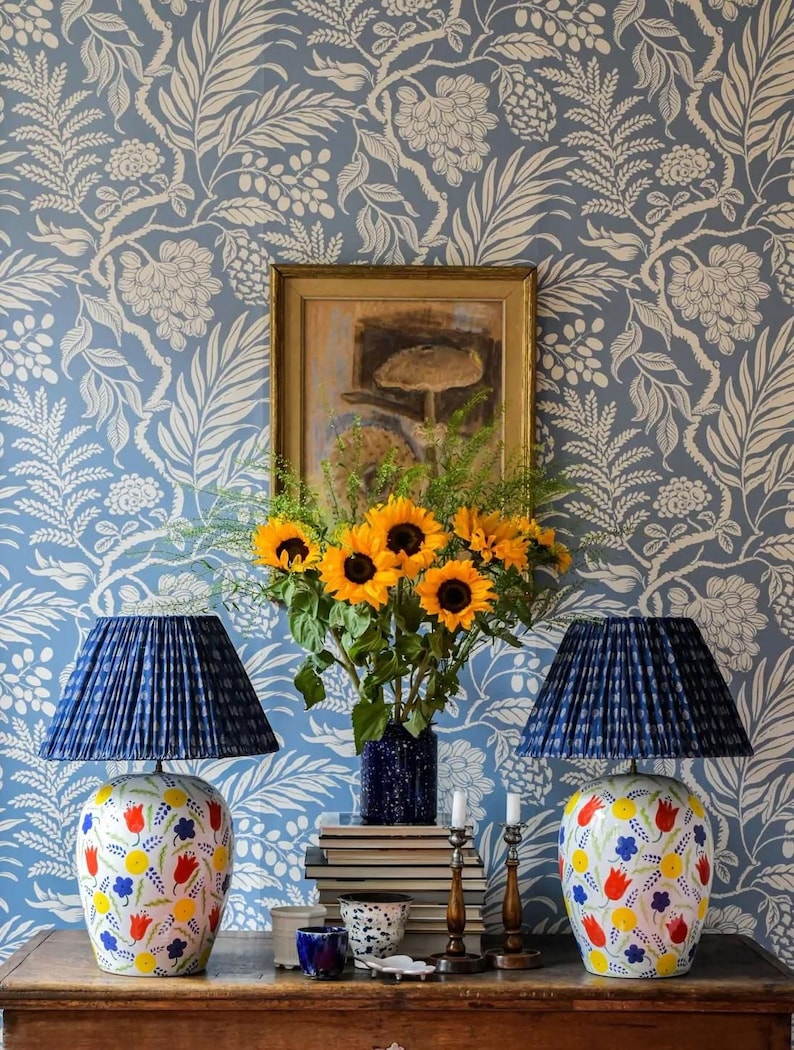 a table with two blue lamps, a vase of yellow sunflowers, and a framed picture on the wall. The table also has a stack of books and some decorative items.