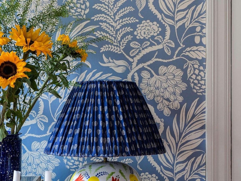 a blue lamp shade with a white pattern, placed on a table next to a vase filled with yellow sunflowers. The background has a blue and white floral wallpaper.