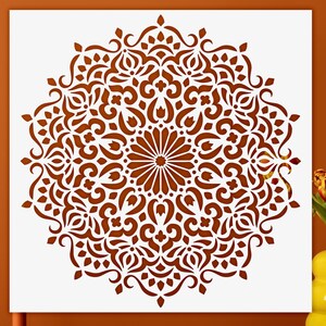 May include: A square white stencil featuring a detailed, symmetrical mandala pattern in a deep rust-orange color. The intricate design consists of radiating floral and geometric shapes, centered around a starburst motif with curved, ornate lines.
