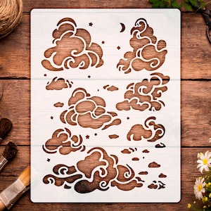 May include: A white stencil featuring cloud designs, stars, and a crescent moon. The stencil is made of a durable material and is ideal for various craft projects. The image is set against a wooden background.
