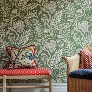 a room with a green floral wallpaper, a red bench with a floral patterned cushion, a wicker chair with a red and white patterned cushion, and a pair of white shoes on the floor.