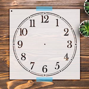 May include: A square stencil featuring a circular clock face design with black Arabic numerals from 1 to 12. The stencil has a small circular hole in the top left corner and a central point for clock mechanism placement, presented on a flat surface.
