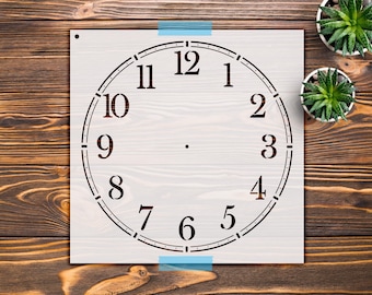 May include: A square stencil featuring a circular clock face design with black Arabic numerals from 1 to 12. The stencil has a small circular hole in the top left corner and a central point for clock mechanism placement, presented on a flat surface.