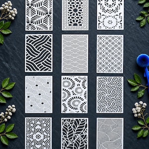 May include: A set of twelve rectangular white stencils featuring various geometric, floral, and abstract patterns, including leaf motifs, wave designs, star-like shapes, and intricate lattice work, arranged in a grid on a dark textured surface.