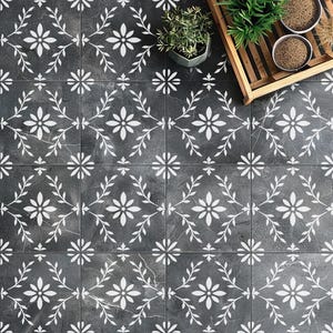 May include: A set of square floor tiles featuring a dark charcoal gray stone texture with a repeating white floral and vine pattern. The tiles are arranged in a grid, creating a continuous botanical design across the surface of the flooring.